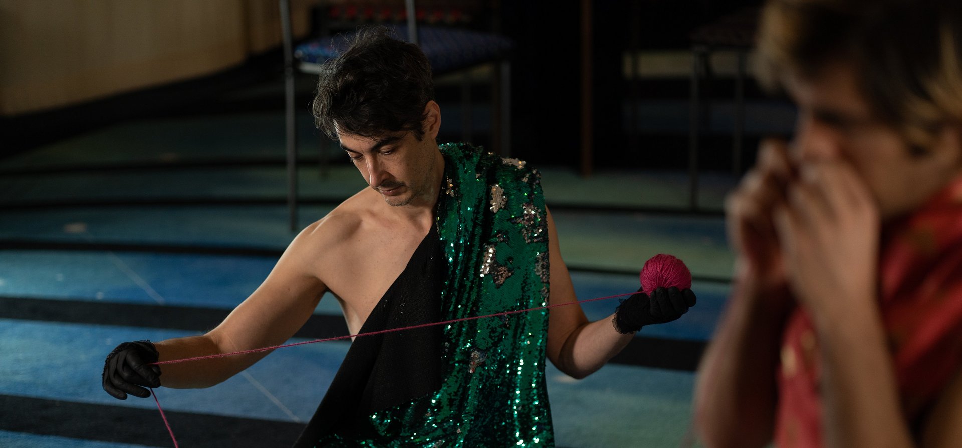 Eine Person in grünem Pailletten-Umhang sitzt mit Wollknäuel am Boden.