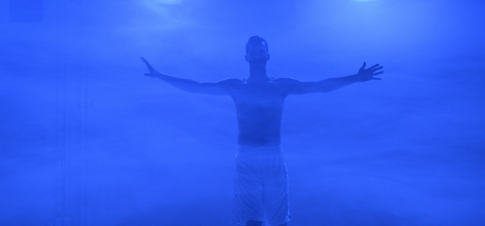 In der Mitte des Bildes steht eine Person mit ausgestreckten Armen. Die Bühne ist in blaues Licht und Nebel gehüllt.