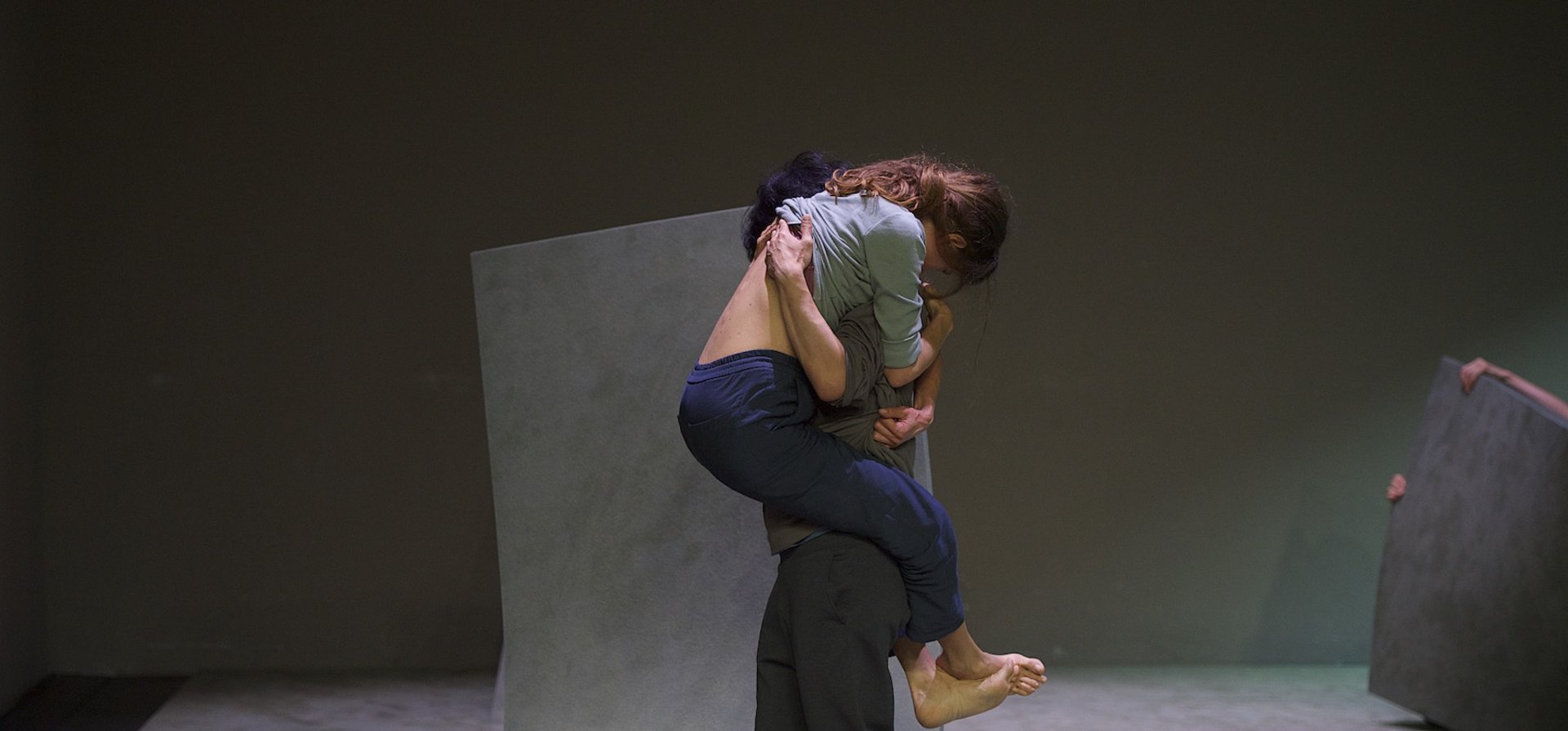 Photography of a stage scene. A person clings to another person. She is being carried by her. Large elastic floor panels are lifted from the floor.