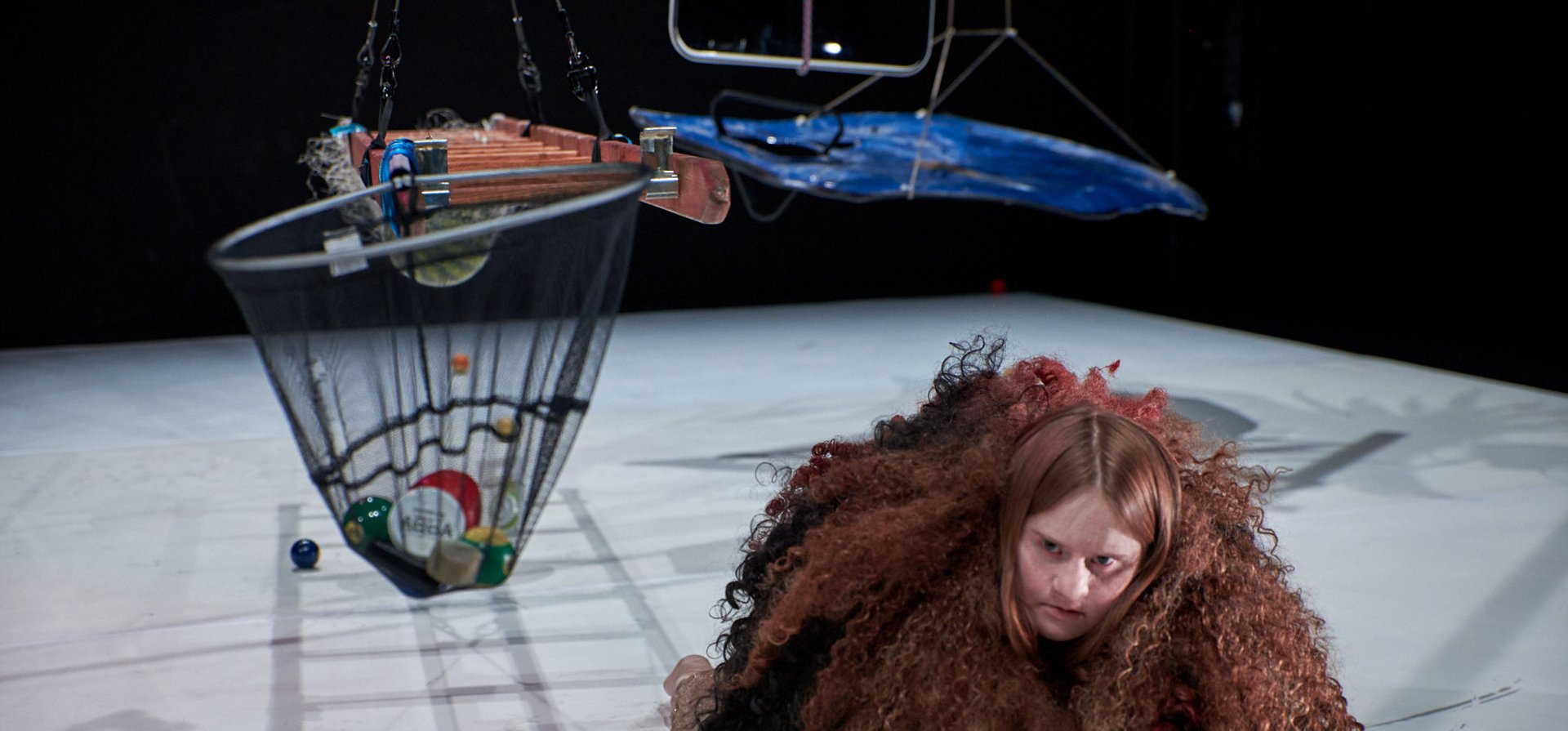 Eine Person kriecht auf allen Vieren auf dem Boden. Sie trägt einen Poncho aus langer lockiger Wolle. Um sie herum hängen ein Flugobjekt, eine Leiter und ein Netz, gefüllt mit Billardkugeln, von der Decke.