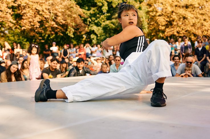 	 Eine Tänzerin in weißen Hosen und einem schwarzen Oberteil tritt auf einer Freilichtbühne auf, umgeben von einer Zuschauermenge. Im Hintergrund sind Bäume mit gelben und grünen Blättern zu sehen.