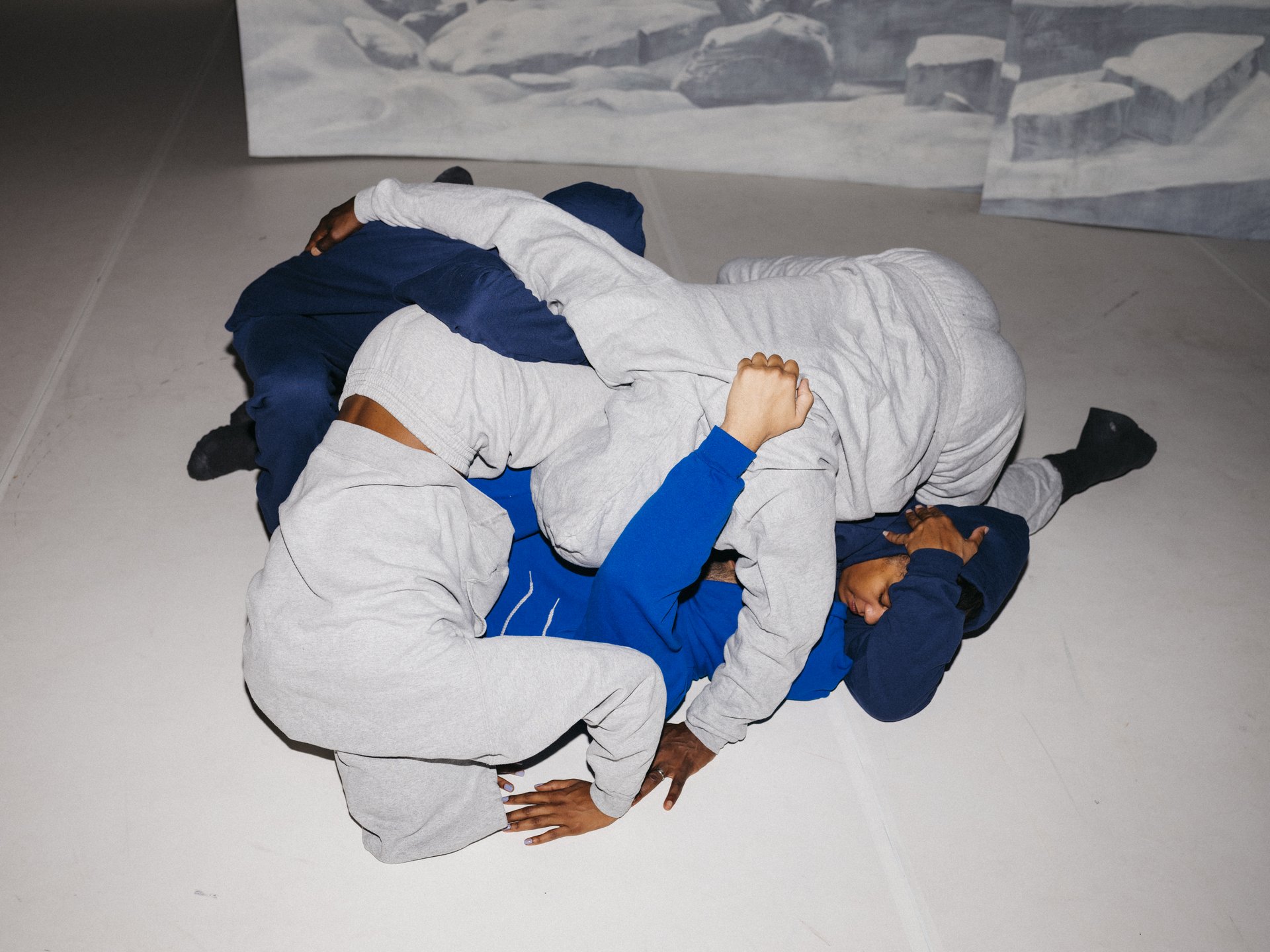 Mehrere Performer:innen in grauen und blauen Trainingsanzügen liegen ineinander verschlungen auf dem Boden vor einer Kulisse mit Eislandschaft.