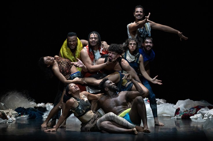 Zehn Performer:innen posieren dicht gedrängt auf einer dunklen Bühne, umgeben von verstreuten Stoffen und weißen Plastikplanen.