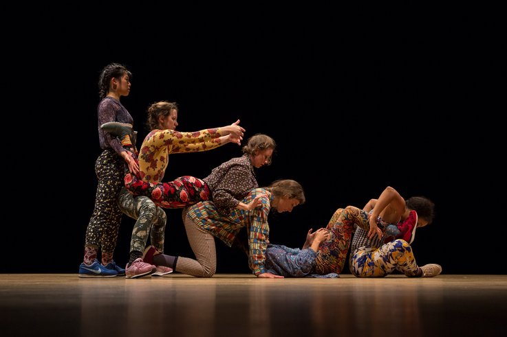 Sieben Performer:innen in gemusterter Kleidung bilden auf der Bühne eine bewegte, skulpturale Menschenformation mit ineinandergreifenden Körpern.
