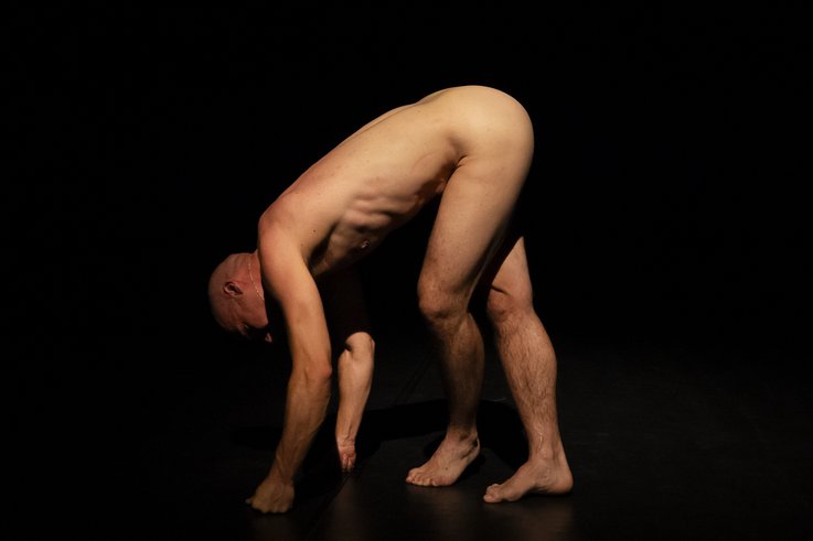 Daniel Mariblanca ist nackt vor einem schwarzen Hintergrund von der Seite fotografiert. Daniel Mariblanca stützt sich mit Händen und Füßen auf dem Boden ab. 