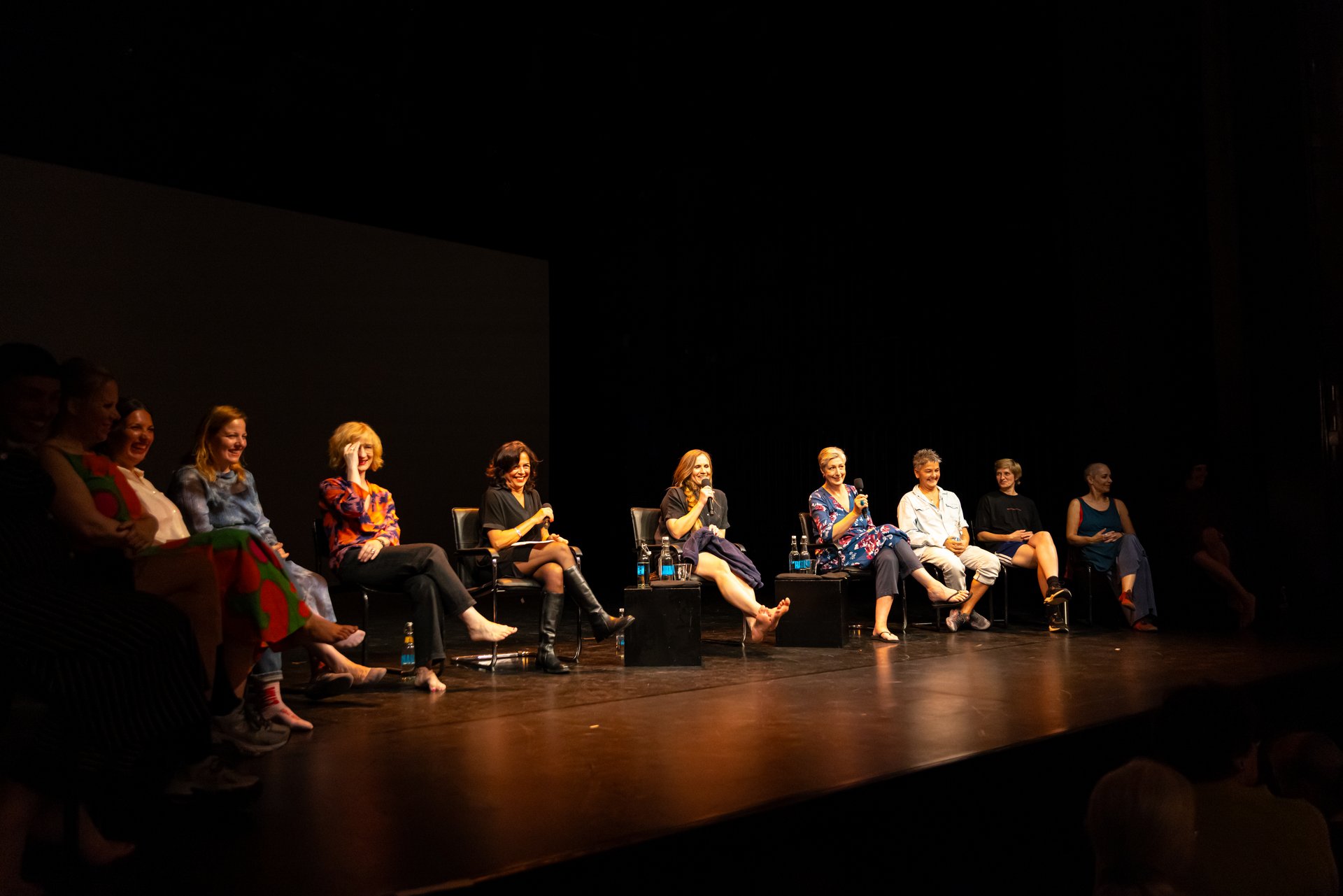 Auf dem Bild sind 10 Personen während des Beyond the Stage mit Canan Erek zu sehen. Die Bühne ist dunkel und alle Frauen sitzen in einer Reihe. In der Mitte ist Lovísa Ósk Gunnarsdóttir zu sehen.