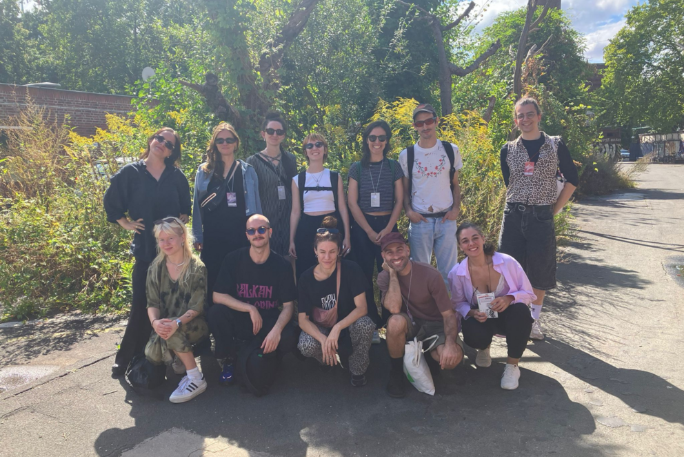 Auf dem Foto sind 12 Personen zu sehen, die sich im Freien für ein Gruppenfoto versammelt haben. Im Hintergrund sind Bäume, ein Stück blauer Himmel und eine kleine Straße zu sehen. 