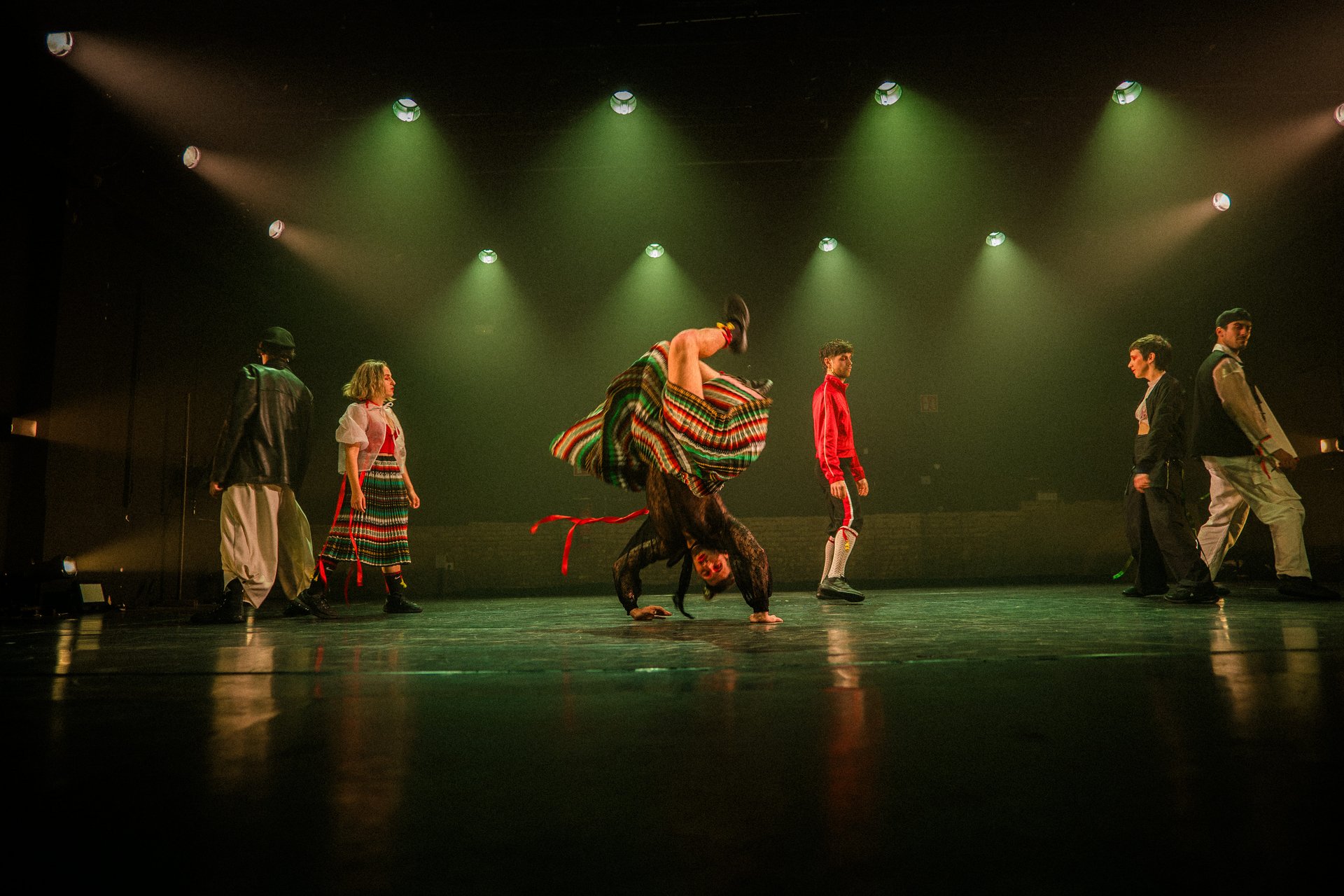 Fünf Performer:innen auf grün beleuchteter Bühne, teils in folkloristischer, teils in sportlicher Kleidung, eine Person im Handstand mit schwingendem Rock.