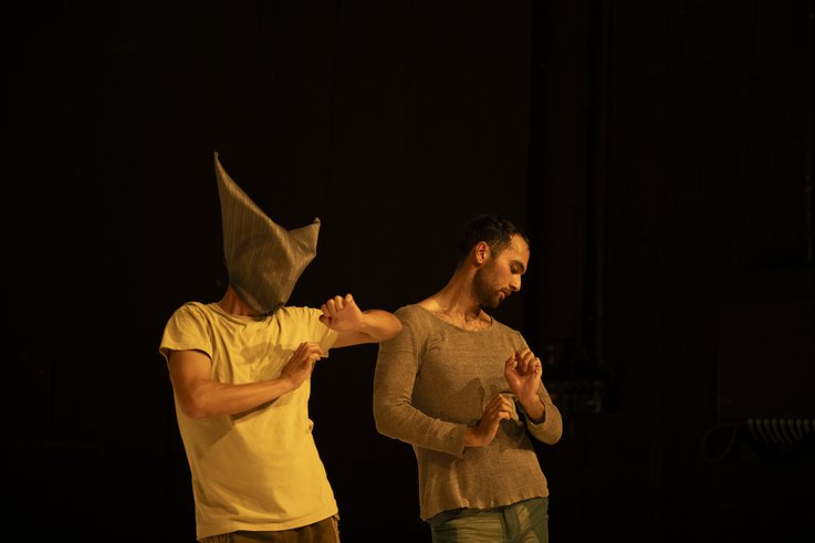 Zwei Tänzer*innen der Compagnie sind als Halbfigur vor einem dunklen Hintergrund fotografiert. Die Person rechts trägt eine Maske und stützt ihren Ellbogen auf die Schulter des anderen.