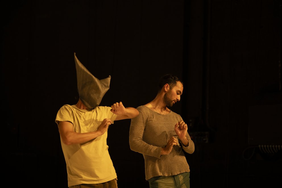 Zwei Tänzer*innen der Compagnie sind als Halbfigur vor einem dunklen Hintergrund fotografiert. Die Person rechts trägt eine Maske und stützt ihren Ellbogen auf die Schulter des anderen.