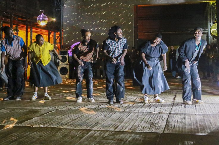 Eine Gruppe von Menschen tanzt energisch in einem großen Veranstaltungsort im Industriestil, der von bunten Lichtern und Discokugeln beleuchtet wird, mit Zuschauern im Hintergrund.