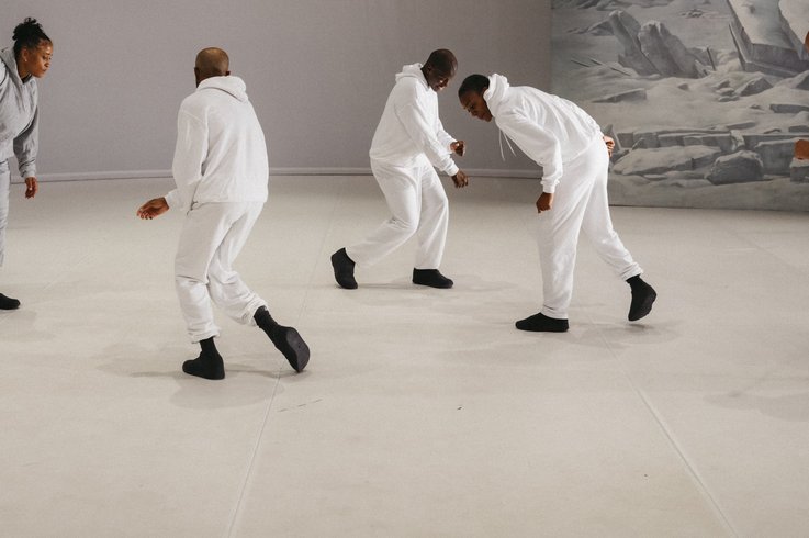 Fünf Performer:innen in weißen und grauen Trainingsanzügen bewegen sich in gebeugter Haltung vor winterlicher Bühnenkulisse.