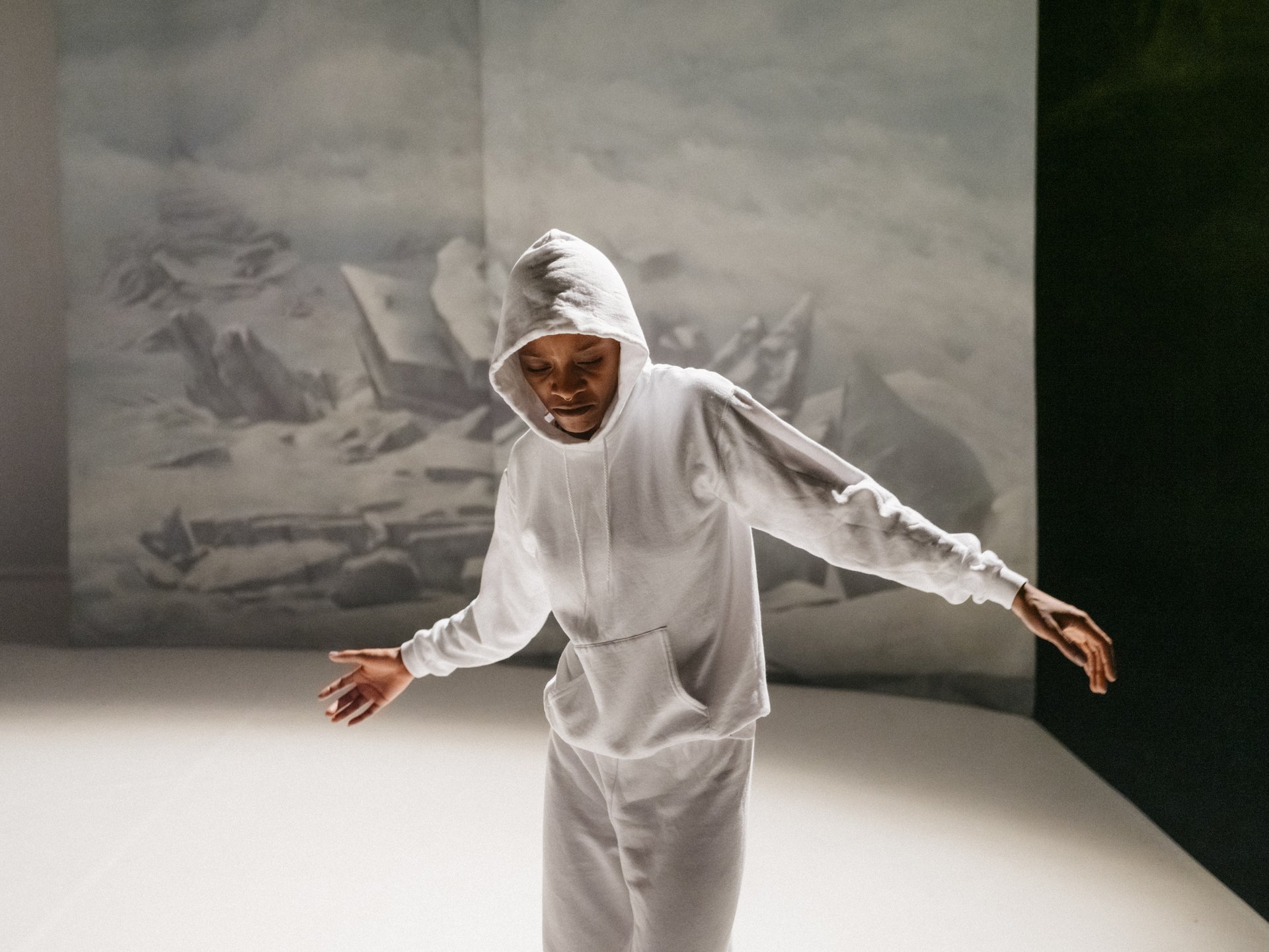 Performer:in in weißem Hoodie und Jogginghose mit ausgestreckten Armen vor gemalter Eislandschaftskulisse auf heller Bühne.