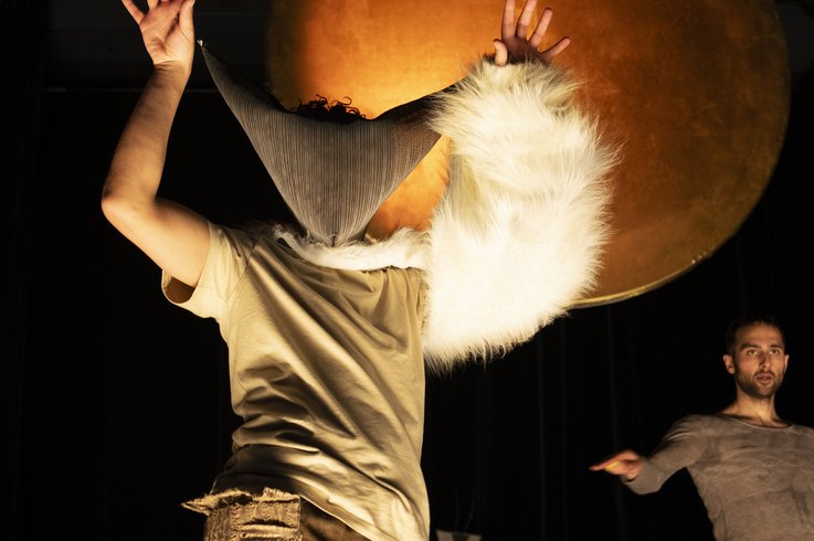 Eine Person mit spitzer Mütze und weißem Pelzärmel steht mit erhobenen Armen vor einer großen, orange beleuchteten Kugel; im Hintergrund ist eine weitere Person mit neutralem Gesichtsausdruck zu sehen.