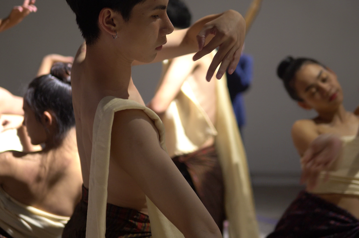 Mehrere Performer:innen mit nacktem Oberkörper und traditionell anmutenden Stoffbahnen in sanften, fließenden Bewegungen; Fokus auf eine zentrale Person mit erhobenen Armen in meditativer Pose.