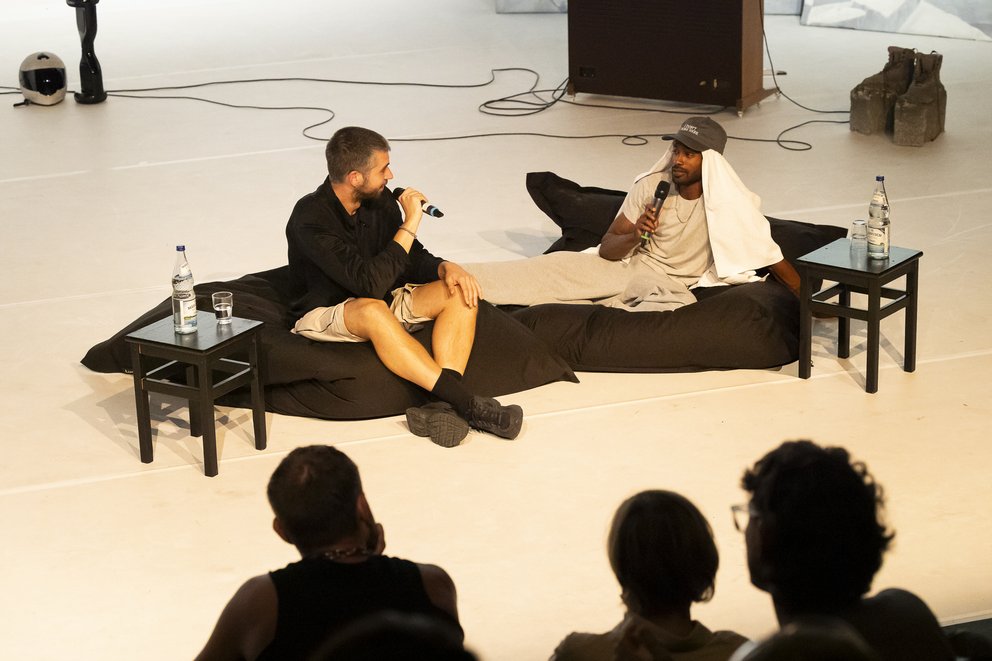 Auf dem Bild ist Beyond the Stage mit Jeremy Nedd und Mateusz Szymanówka zu sehen. Die beiden sitzen auf zwei Poufs und schauen sich an.