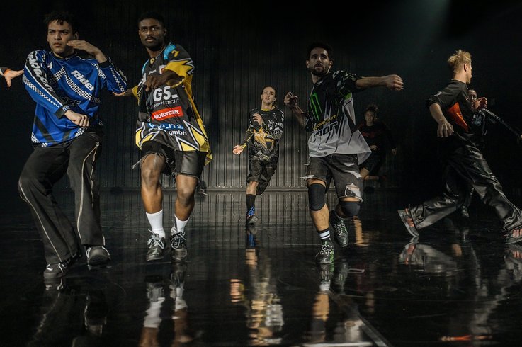 Eine Gruppe von fünf Männern tanzt energisch auf einer dunklen, glänzenden Bühne. Sie tragen bunte, übergroße Sporttrikots und -hosen und werden von einer dramatischen Beleuchtung von oben angestrahlt.