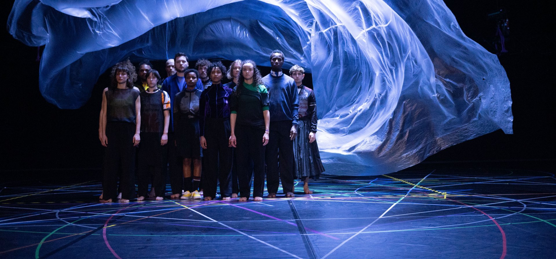 a group of dancers are standing in a black space under a bluely illuminated floating plastic that floats above their heads