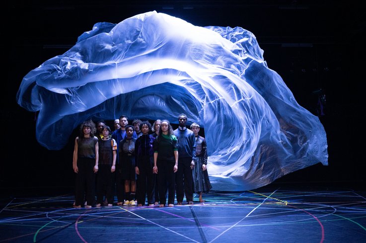a group of dancers are standing in a black space under a bluely illuminated floating plastic that floats above their heads