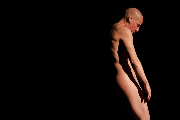 Eine nackte Person mit rasiertem Kopf steht seitlich im dramatischen Bühnenlicht, der Körper in sich versunken, vor schwarzem Hintergrund.