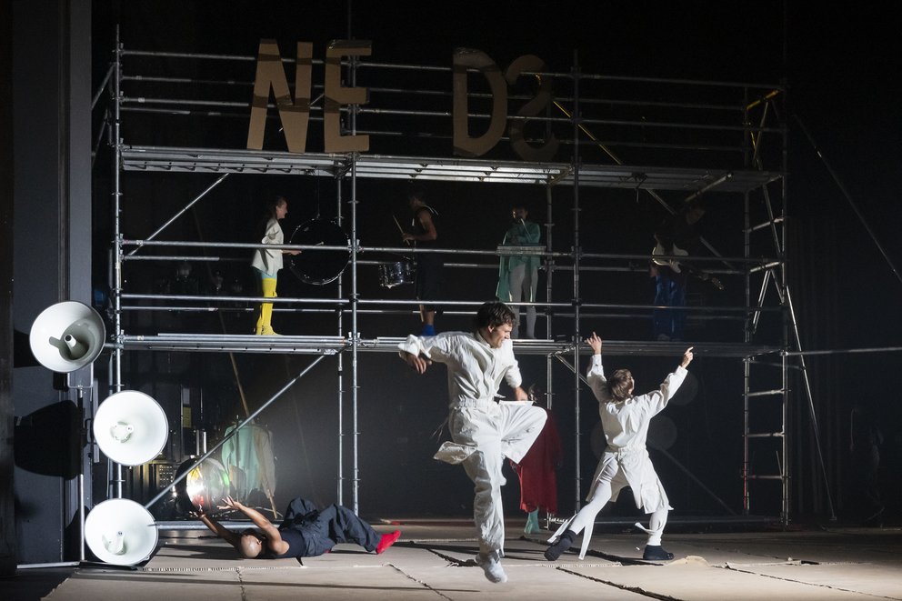 Auf der Bühne sieht man das Bühnenbild der Produktion, bestehend aus einer Metallkonstruktion im Hintergrund. Davor tanzen zwei Schauspieler*innen in weißen Overalls.