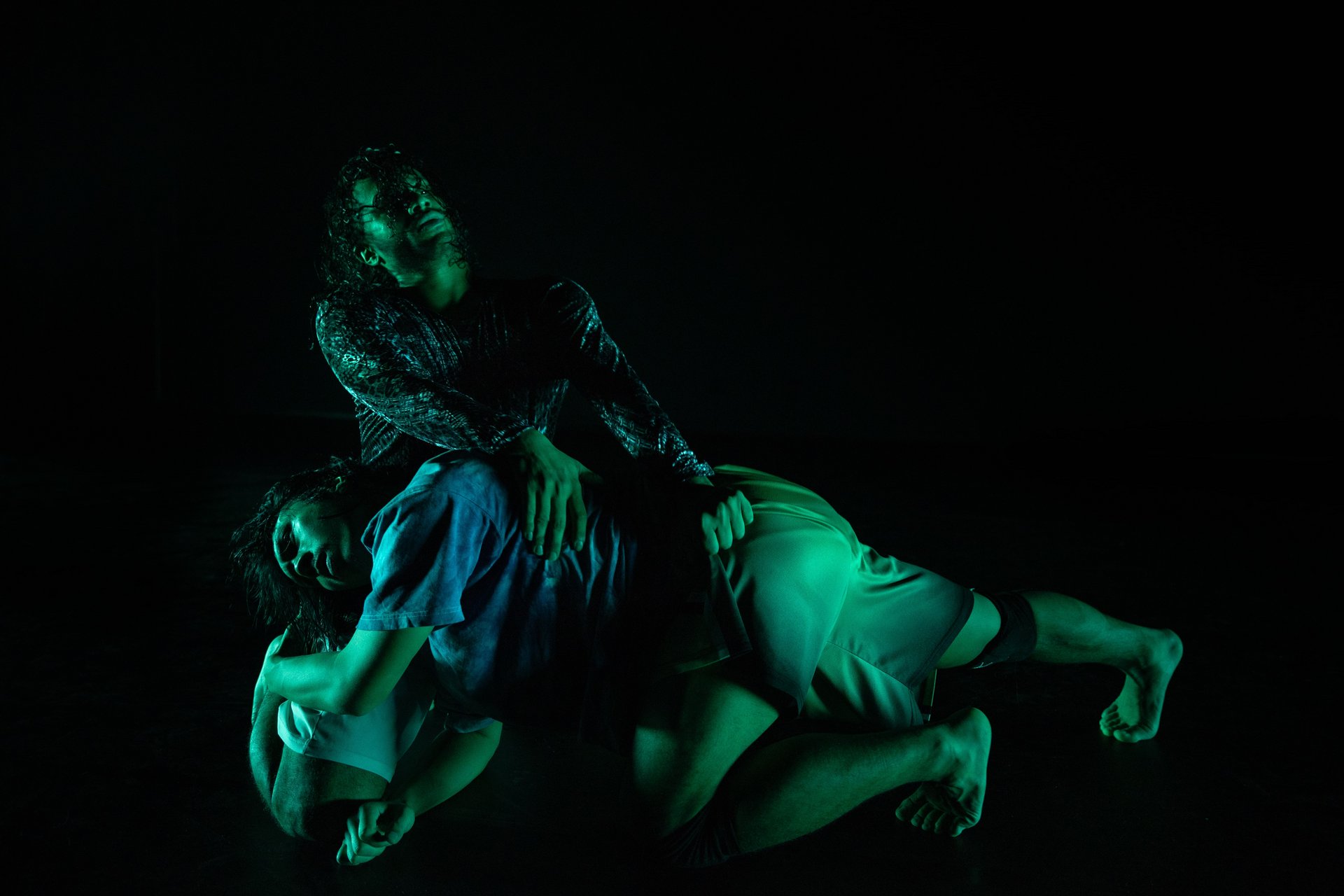 Zwei Performer:innen in dramatischer, körperlich verschränkter Pose auf dunkler Bühne, grünlich beleuchtet; eine Person liegt über der anderen und blickt nach oben.