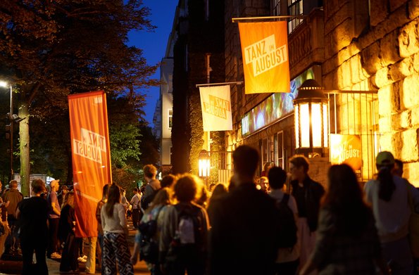 Auf dem Bild sind Menschen vor dem HAU1 nach der letzten Aufführung des Festivals zu sehen. Die Abendbeleuchtung ist eingeschaltet.