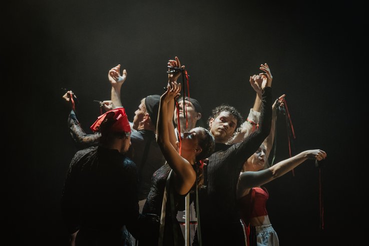 Acht Performer:innen stehen eng beisammen, die Arme anmutig erhoben, rote und grüne Bänder in den Händen – ein kraftvoller, ritueller Moment in dramatischem Bühnenlicht.