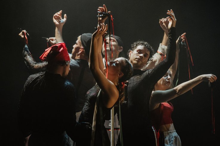 Acht Performer:innen stehen eng beisammen, die Arme anmutig erhoben, rote und grüne Bänder in den Händen – ein kraftvoller, ritueller Moment in dramatischem Bühnenlicht.