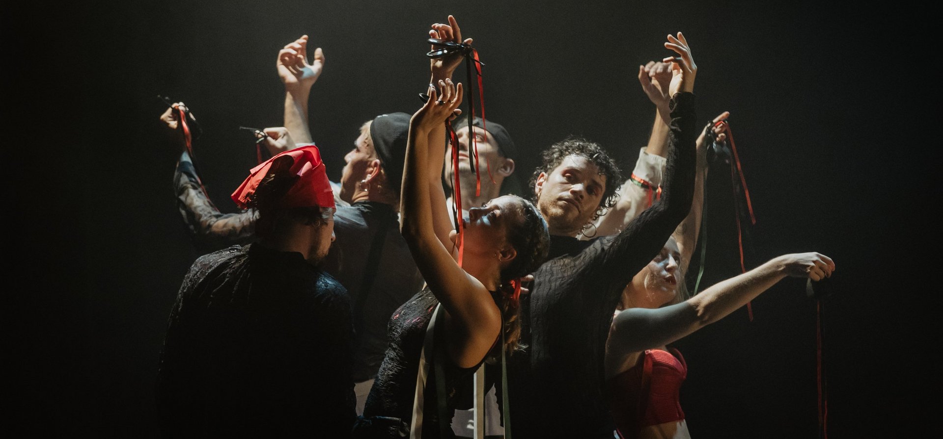 Acht Performer:innen stehen eng beisammen, die Arme anmutig erhoben, rote und grüne Bänder in den Händen – ein kraftvoller, ritueller Moment in dramatischem Bühnenlicht.