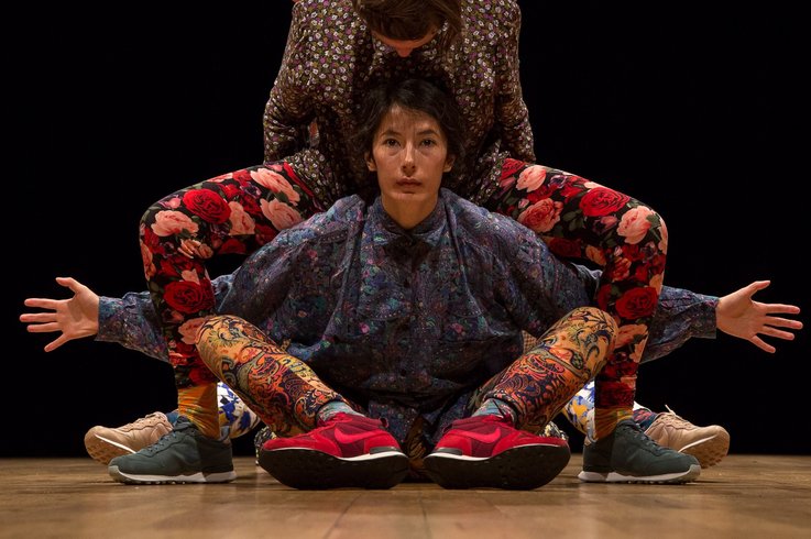 Drei Performer:innen in farbenfrohen, floralen und ornamentalen Outfits verschmelzen zu einer symmetrischen Figur auf der Bühne, vor schwarzem Hintergrund.