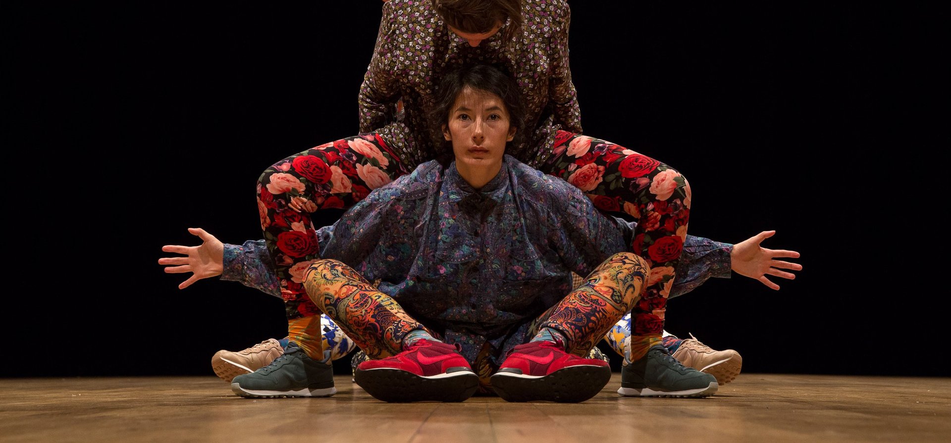 Drei Performer:innen in farbenfrohen, floralen und ornamentalen Outfits verschmelzen zu einer symmetrischen Figur auf der Bühne, vor schwarzem Hintergrund.