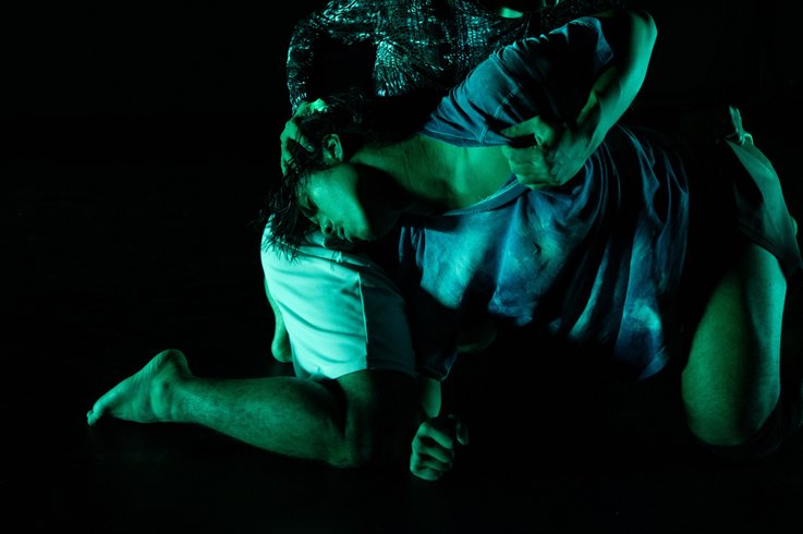 Zwei Performer:innen in intensiver Pose auf dunkler Bühne, grünlich beleuchtet, mit ineinander verschlungenen Körpern.