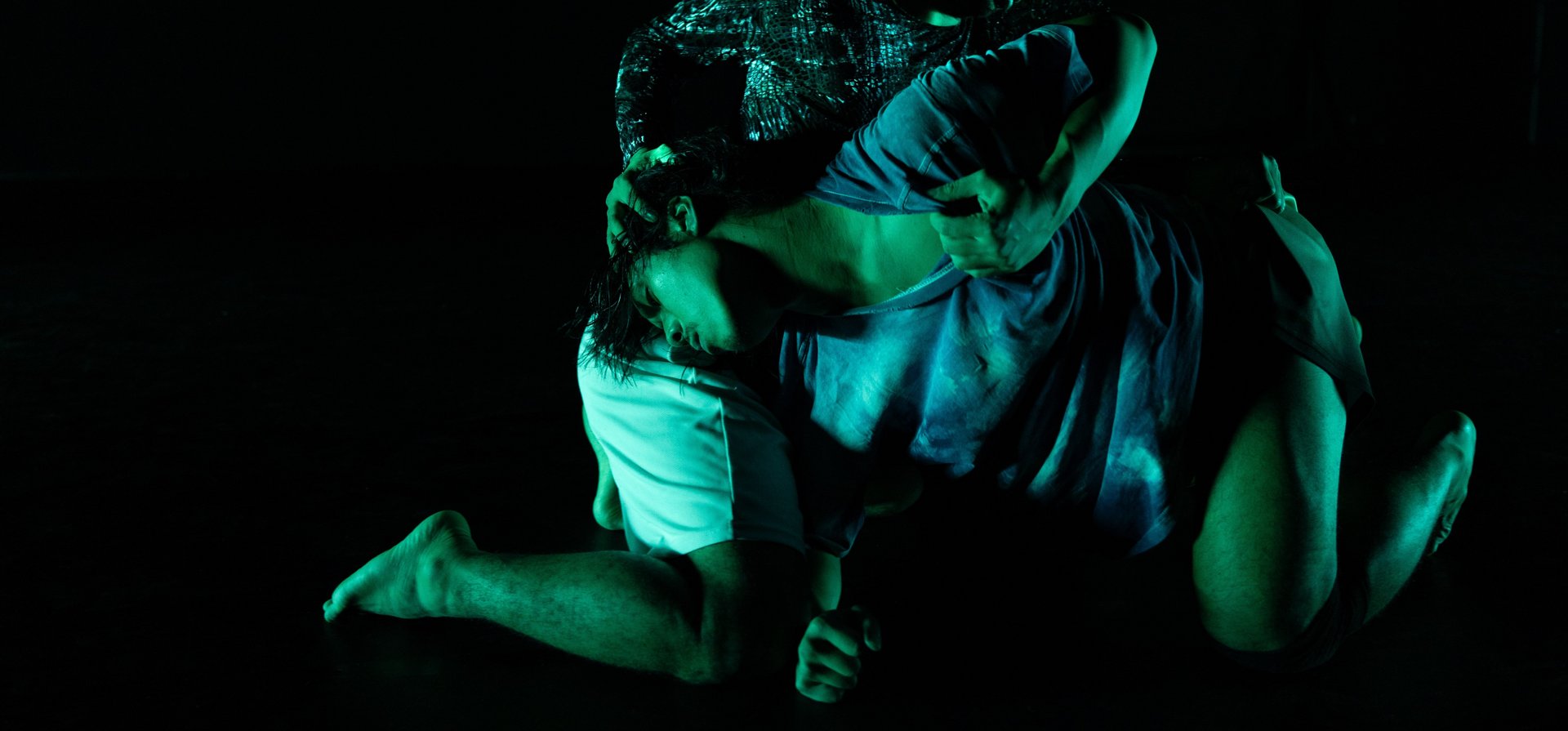 Zwei Performer:innen in intensiver Pose auf dunkler Bühne, grünlich beleuchtet, mit ineinander verschlungenen Körpern.