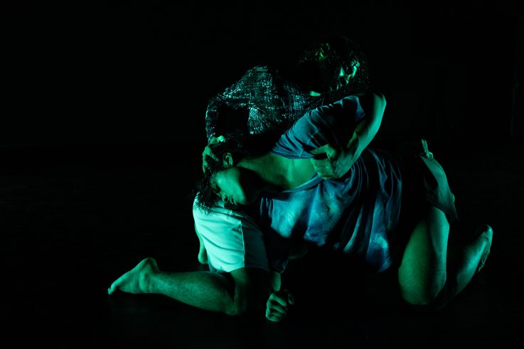Zwei Performer:innen in intensiver Pose auf dunkler Bühne, grünlich beleuchtet, mit ineinander verschlungenen Körpern.