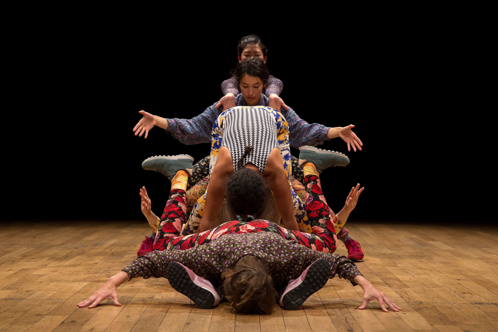 Sechs Performer:innen in farbenfroher Kleidung bilden eine übereinandergestapelte Formation auf der Bühne – eine dynamische, skulpturale Choreografie.