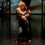 Eine barfüßige Performer:in mit blondem Haar tanzt allein auf dunkler Bühne, im Hintergrund sind großformatige Videoausschnitte mit Innenraumszenen zu sehen.