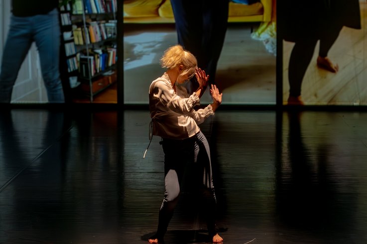 Eine barfüßige Performer:in mit blondem Haar tanzt allein auf dunkler Bühne, im Hintergrund sind großformatige Videoausschnitte mit Innenraumszenen zu sehen.
