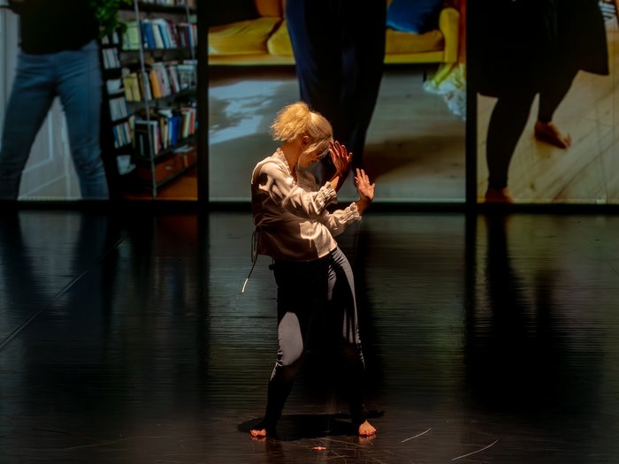 Eine barfüßige Performer:in mit blondem Haar tanzt allein auf dunkler Bühne, im Hintergrund sind großformatige Videoausschnitte mit Innenraumszenen zu sehen.