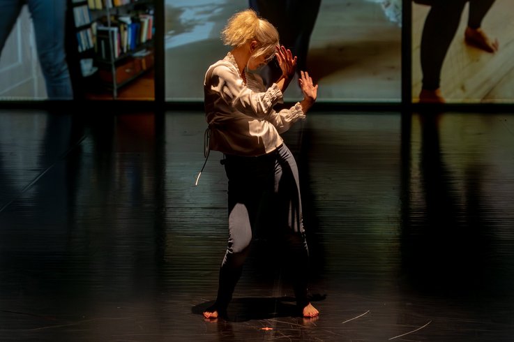 Eine barfüßige Performer:in mit blondem Haar tanzt allein auf dunkler Bühne, im Hintergrund sind großformatige Videoausschnitte mit Innenraumszenen zu sehen.