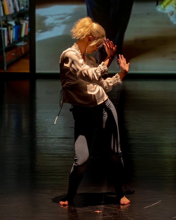 Eine barfüßige Performer:in mit blondem Haar tanzt allein auf dunkler Bühne, im Hintergrund sind großformatige Videoausschnitte mit Innenraumszenen zu sehen.