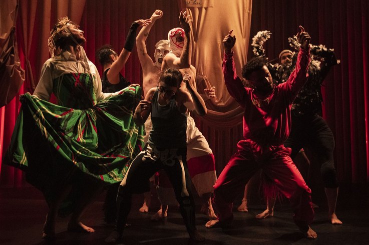 Eine Gruppe von Darstellern in farbenfrohen, dramatischen Kostümen posiert dynamisch auf einer schwach beleuchteten Bühne mit roten Vorhängen und drückt unter der Theaterbeleuchtung intensive Emotionen und Bewegungen aus.