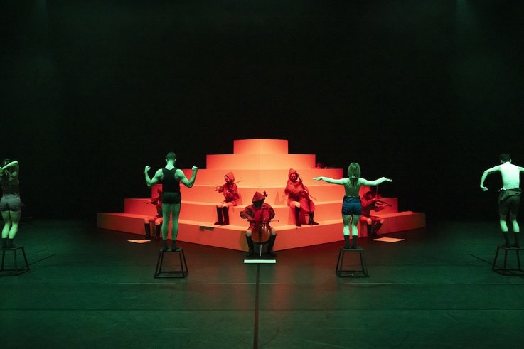 Performer:innen auf Hockern und Musiker:innen in roten Kapuzen posieren vor einer stufenförmigen weißen Plattform im grünen und roten Licht.
