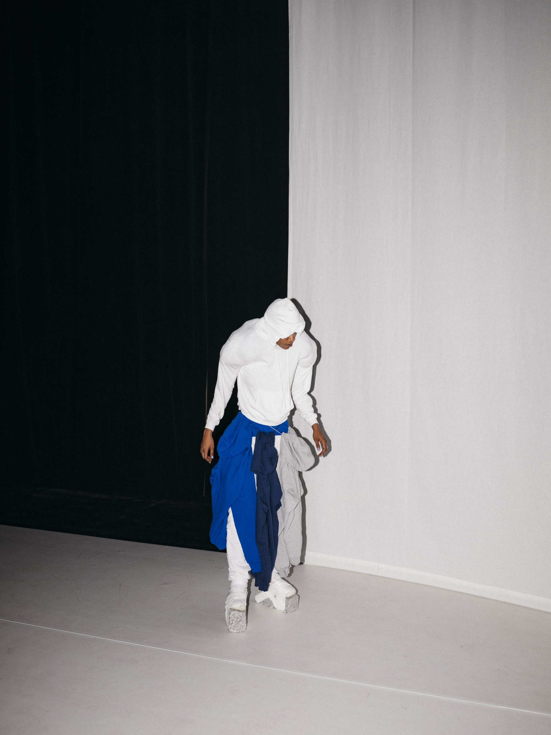 Performer:in in weißem Outfit mit Kapuze und mehreren umgebundenen Pullovern vor kontrastierender weiß-schwarzer Wand auf heller Bühne.