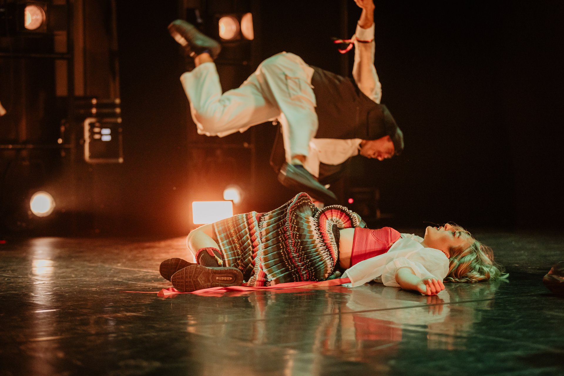 Performer:in in folkloristischer Kleidung liegt am Boden, im Hintergrund springt ein:e Performer:in in weißem Kostüm vor Scheinwerferlicht.