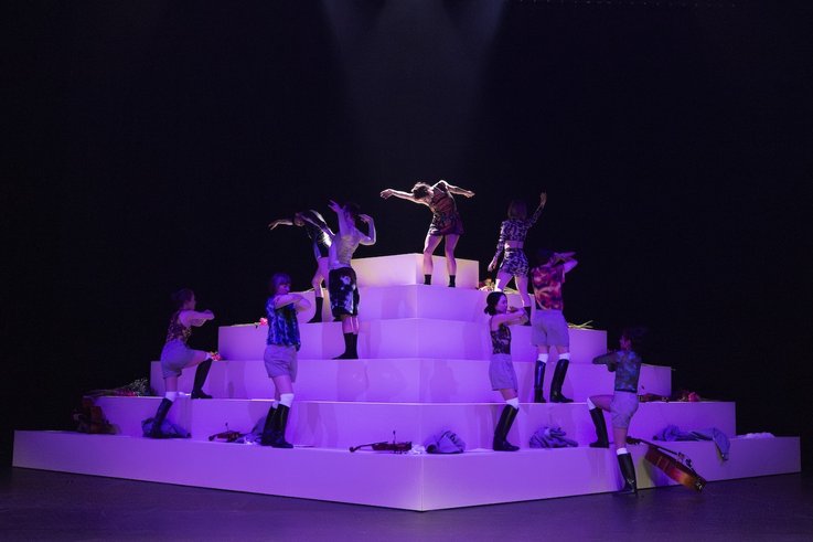 Performer:innen bewegen sich asynchron auf einer stufenförmigen weißen Plattform unter violettem Licht, umgeben von Kleidung und Instrumenten.