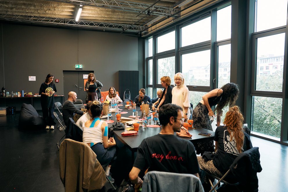 Auf dem Foto sieht man 13 Künstler*innen an einem Tisch, die sich unterhalten und kennenlernen. Im Hintergrund sieht man große Fenster. 