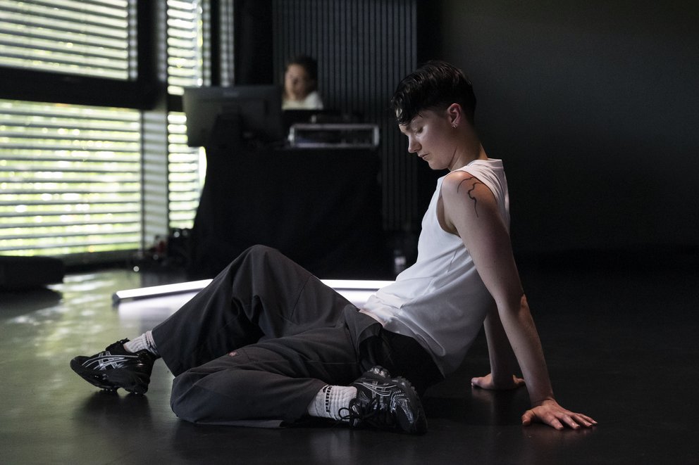 Xan Dye sitzt in der Mitte des Bildes auf dem Boden und schaut zu Boden. Im Hintergrund ist ein Fenster zu sehen.