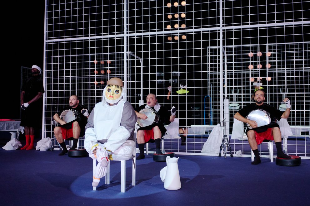 Auf dem Bild sieht man im Hintergrund eine Struktur aus weißen Eisengittern, im Vordergrund eine*n Schauspieler*in, der/die eine Puppenmaske trägt. Dahinter sind auch andere sitzende Schauspieler*innen zu erkennen.