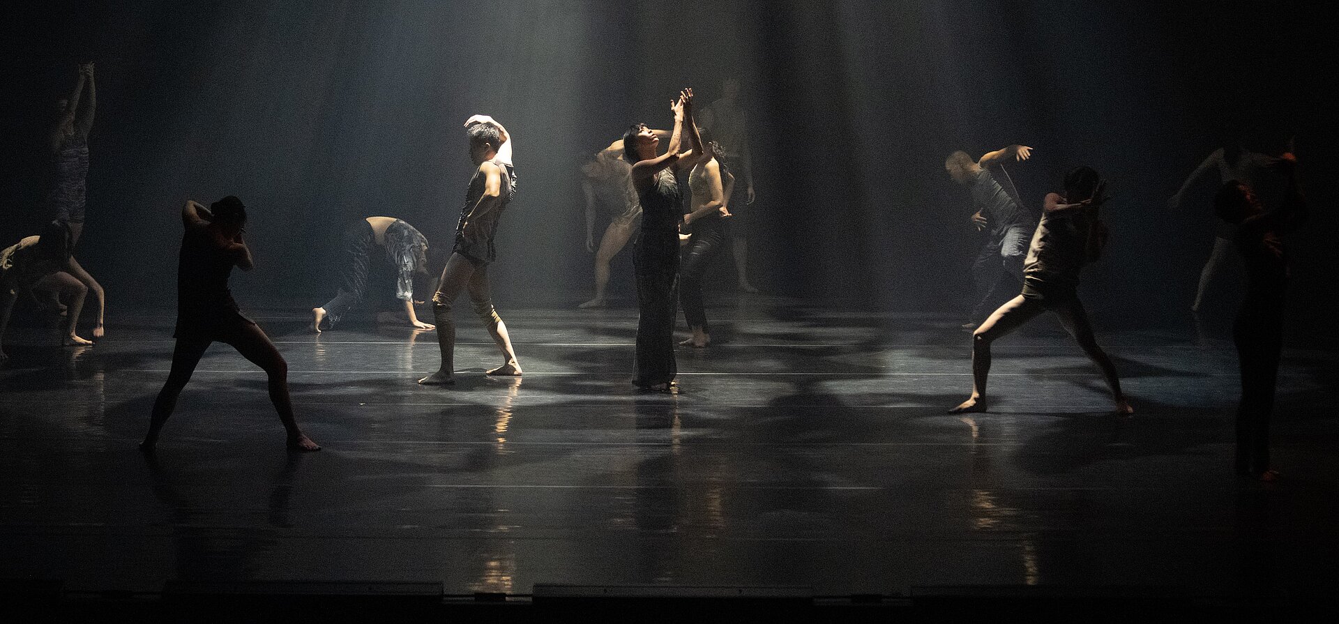 Gruppe von Performer:innen auf dunkler Bühne, die sich in verschiedenen Posen bewegen; von oben fällt ein gebündelter Lichtstrahl auf die Szene.